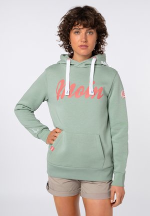 Frau mit lockigem Haar trägt einen hellgrünen Hoodie mit pinkem Schriftzug „Moin“, beige Shorts, eine Hand in der Hüfte, stehend vor neutralem Hintergrund.
