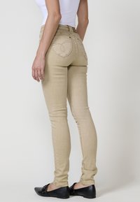 Vaqueros skinny beige con un diseño ajustado, que presentan costuras decorativas en los bolsillos traseros, combinados con zapatos planos negros. Textura suave.