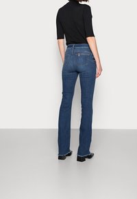 Kobieta nosząca niebieskie dżinsy z wysokim stanem i postrzępionymi szwami oraz czarną bluzkę z krótkim rękawem, stojąca na tle jednolitego białego tła.