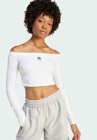 Biely crop top s odhalenými ramenami a rebrovanou textúrou, so malým čiernym logom Adidas. Kombinovaný s svetlošedými nohavicami s elastickým pásom.