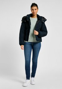 Giacca a vento nera con cappuccio, chiusura a zip e dettagli in bottoni. Presenta un colletto alto e polsini a costine. Abbinata a un maglione verde chiaro e jeans blu.