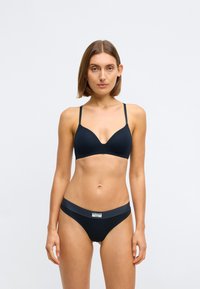 Femme au short cheveux bruns portant un soutien-gorge noir et une culotte noire assortie avec une étiquette sur la taille, se tenant devant un fond clair.