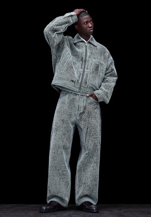 Jonge man poseert in een gestructureerde, oversized grijze jas en wijde broek met zwarte schoenen tegen een zwarte achtergrond.