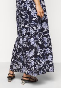 Kvinna i en marinblå blommig kjol och svarta klacksandaler, stående med handen avslappnat vid sin sida.