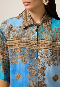 Cămașă cu nasturi, având un model paisley albastru și maro, cu guler structurat și mâneci scurte, fabricată dintr-un material ușor.