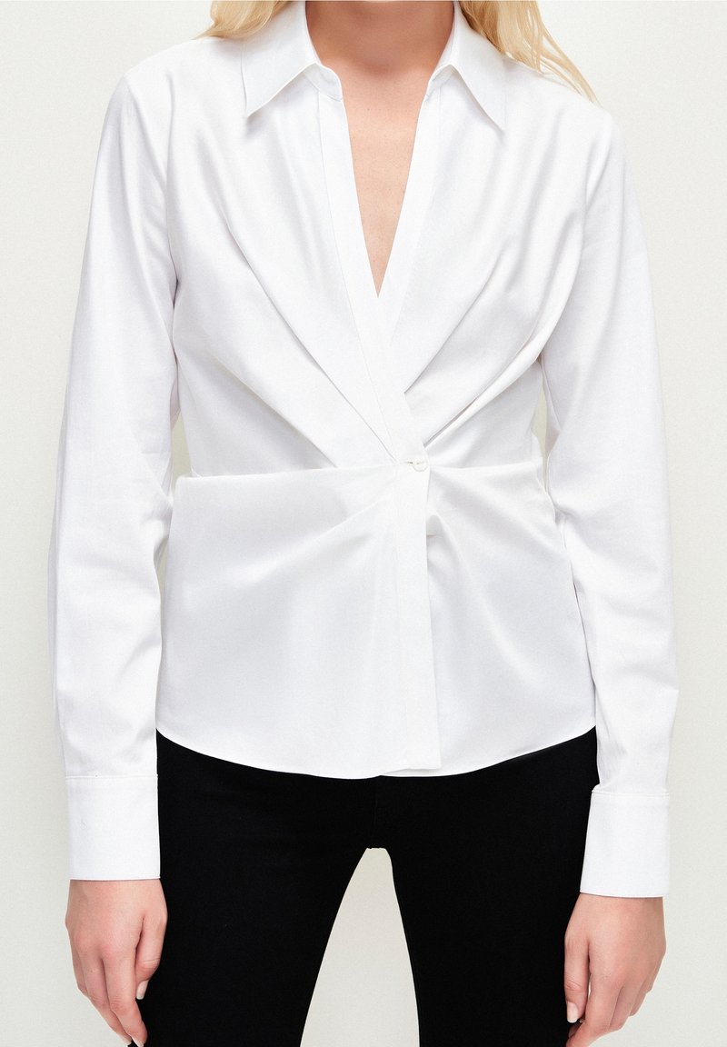 Chemise blanche à manches longues avec un profond décolleté en V, un design noué sur le devant, une fermeture par un seul bouton, et un tissu en coton lisse. Associée à un pantalon noir.