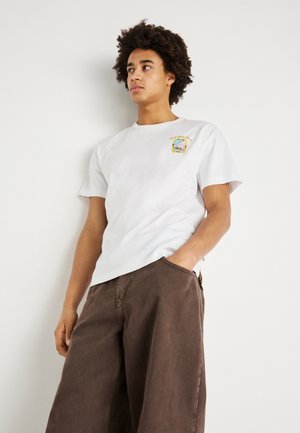 Camiseta blanca de algodón con mangas cortas y cuello redondo, que presenta un pequeño gráfico colorido en el lado izquierdo del pecho. Pantalones marrones de pierna ancha.