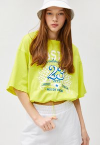 Neon grøn oversized t-shirt med hvide og blå grafikker, der viser tekst og et ternet mønster, båret med elastiske hvide shorts.