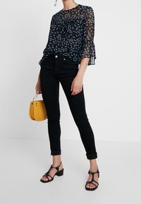 Schwarze, durchsichtige Bluse mit blau gepunktetem Muster und ausgestellt geschnittenen Ärmeln, kombiniert mit schwarzen Skinny Jeans und schwarzen Riemchenheels, während sie eine gelbe Handtasche hält.