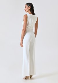 Jumpsuit blanc sans manches avec un corsage ajusté et un pantalon à jambe évasée. Tissu lisse et détail de fermeture éclair au dos. Porté avec des talons à lanières.