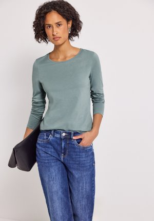 Vrouw met krullend haar, gekleed in een groen shirt met lange mouwen en blauwe jeans, die met één hand een opgevouwen zwart jasje vasthoudt.