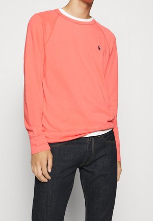Felpa a maniche lunghe color corallo con scollatura a girocollo e polsini a costine, con un piccolo logo vicino al petto, abbinata a jeans scuri.