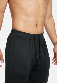 Svarta joggingbyxor tillverkade av mjukt material, med ett elastiskt midjeband och dragband. Sidofickor synliga på varje sida.