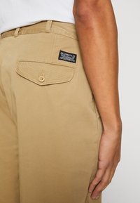 Desni bočni pogled na osebo, ki nosi bež hlače Levi Strauss s gumbom na zadnjem žepu in vidno oznako Levi's.