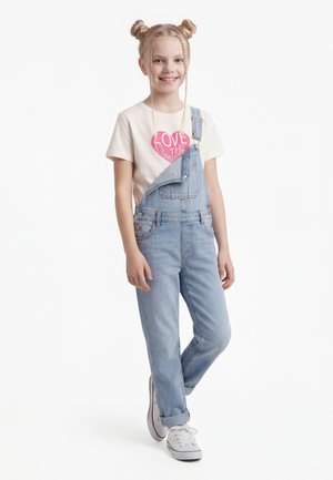 Nasmejana deklica z dvema figama, oblečena v majico z napisom "Ljubezen je odgovor", zavihane denim hlače na naramnice in bele superge.