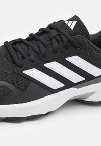 Chaussure de sport noire en matériau en maille, avec un design à trois bandes blanches, une semelle extérieure texturée et des accents gris. Équipée de lacets noirs et d'un col rembourré.