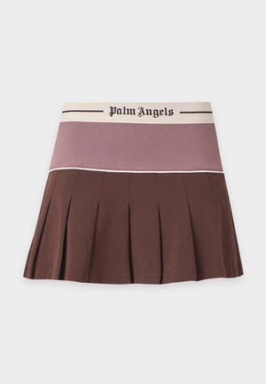 Frau trägt ein braunes, kurz geschnittenes Poloshirt und einen plissierten Minirock mit "Palm Angels"-Branding, steht vor einem schlichten hellen Hintergrund.