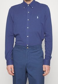 Man som bär en blå långärmad skjorta med knappar och en liten vit logotyp på vänster bröst, i kombination med blå byxor med elastisk midja.