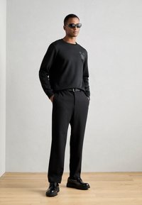 Sudadera negra con detalle estampado en el pecho, pantalones negros y zapatos negros. El modelo también lleva gafas de sol negras y está de pie contra un fondo neutro.