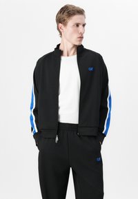 Veste zippée noire avec des rayures bleues sur les manches, dotée d'un col montant et d'un logo. Assortie à un pantalon coordonné, le tout en tissu lisse.