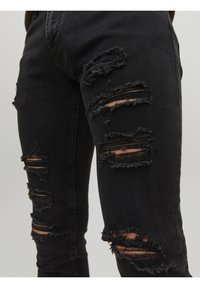 Vaqueros negros desgastados con múltiples desgarros y bordes deshilachados, hechos de denim con un acabado suave y texturizado.
