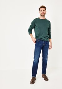 Dunkelgrünes Langarmhemd kombiniert mit blauen Jeans. Braune Lederschuhe runden das Outfit ab. Ein schlichter Hintergrund zeigt die Kleidung.
