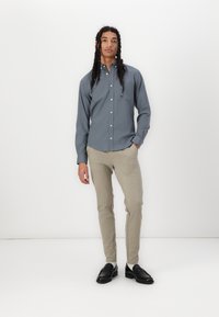 Camisa de botones gris con un bolsillo en el pecho, pantalones beige ajustados y mocasines negros, de pie contra un fondo blanco.