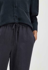 Tummanvihreä nappipaita, jossa on tasku, yhdistettynä löysiin tummanharmaisiin housuihin, joissa on elastinen vyötärö ja nyöri.