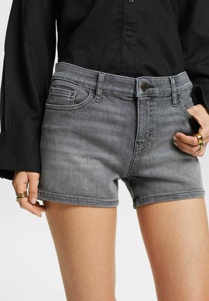 Person iført grå denimshorts og en sort skjorte med knapper, med ringe på begge hænder, den ene hånd i lommen.
