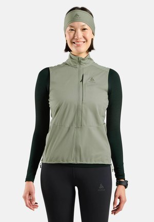 Helles grünes, mit Reißverschluss versehenes Weste mit zwei Fronttaschen, kombiniert mit einem schwarzen Langarmtop mit strukturiertem Muster. Schwarze Leggings und ein Stirnband.