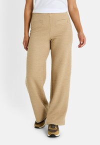 Pantalones de pierna ancha beige, hechos de una tela texturizada, que cuentan con dos bolsillos traseros y un corte recto, combinados con zapatillas deportivas marrones.