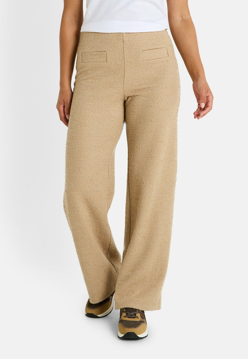 Pantalones de pierna ancha beige, hechos de una tela texturizada, que cuentan con dos bolsillos traseros y un corte recto, combinados con zapatillas deportivas marrones.
