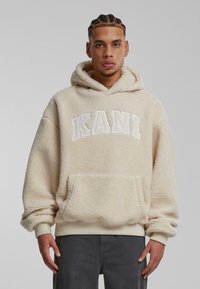 Felpa in pile beige con tasca a marsupio, polsini e orlo ribattuti. La parte anteriore presenta un logo bianco "KANI". Materiale morbido e testurizzato.