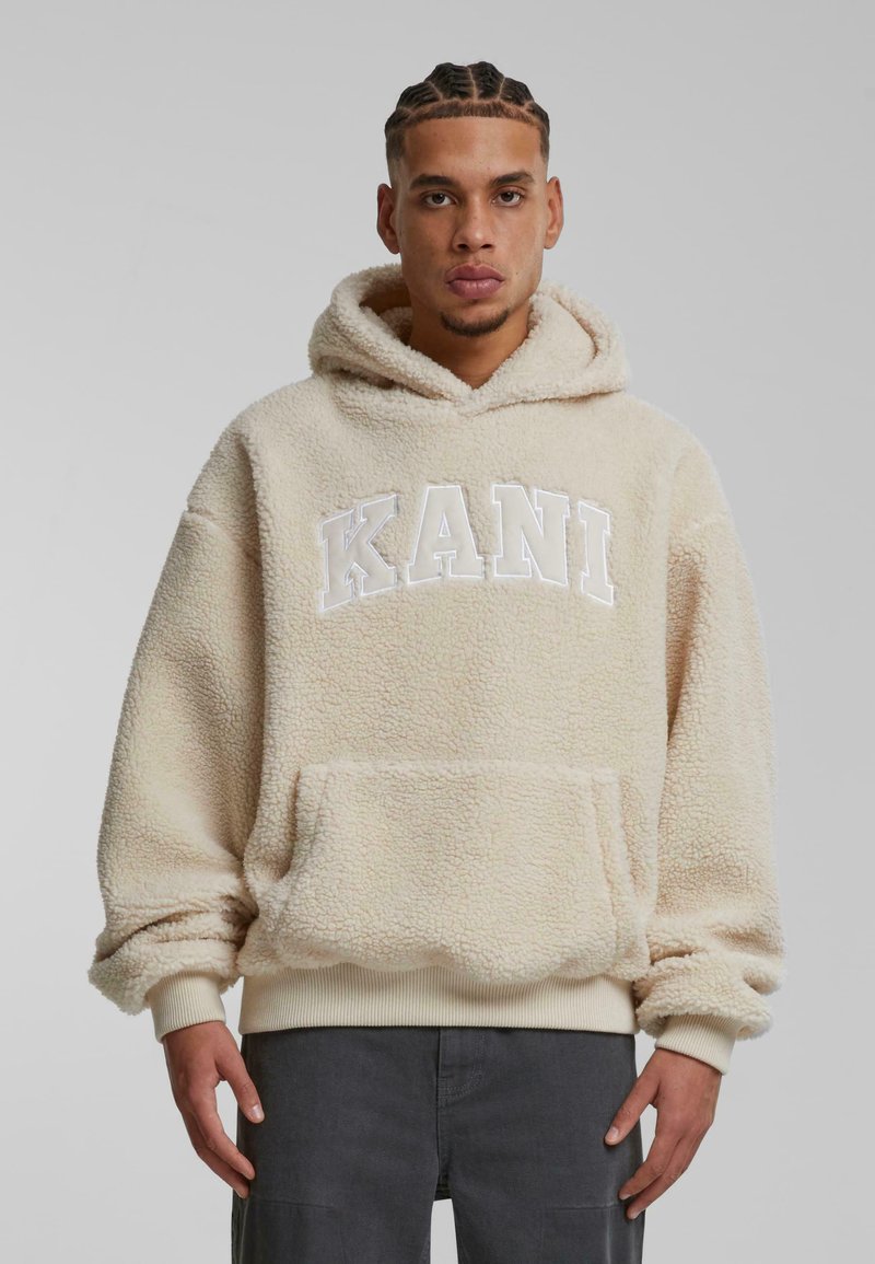 Sudadera de forro polar beige con un bolsillo tipo canguro, puños y dobladillo acanalados. La parte delantera presenta un logotipo en blanco "KANI". Material suave y texturizado.