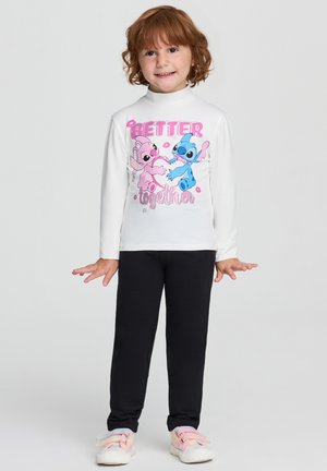 Bambino con capelli ricci indossa una camicia bianca a maniche lunghe con personaggi dei cartoni animati blu e rosa e il testo "Better Together", pantaloni neri e scarpe pastello.