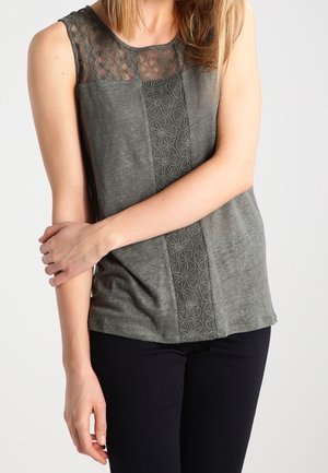 Femme portant un haut gris sans manches avec des détails en dentelle sur la poitrine et au centre, associé à un pantalon noir, debout devant un fond uni.