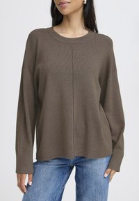 Pull en tricot marron avec un col rond, des épaules tombantes et des poignets côtelés, associé à un denim bleu clair.