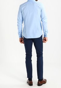 Camicia azzurra a maniche lunghe con un design pulito, polsini con bottoni e bottoni scuri, abbinata a pantaloni slim-fit blu navy e scarpe eleganti marroni.