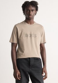 Beige bavlněné tričko s krátkým rukávem, s vyvýšeným logem "BOSS" v mírně tmavší barvě. V kombinaci s černými kalhotami.