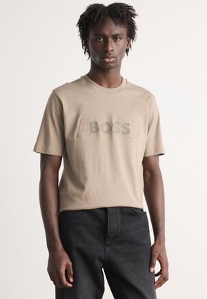 Beige katoenen T-shirt met korte mouwen, met een verhoogd "BOSS" logo in een iets donkere kleur. Gecombineerd met zwarte broeken.