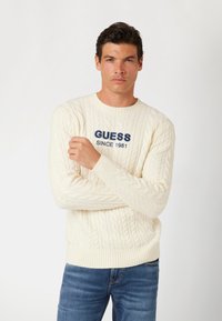 Pulover crem din tricot cu o împletitură fină, realizat dintr-un material moale. Prezintă manșete și tiv cu cusătură în relief, având un logo brodat proeminent albastru "GUESS SINCE 1981."