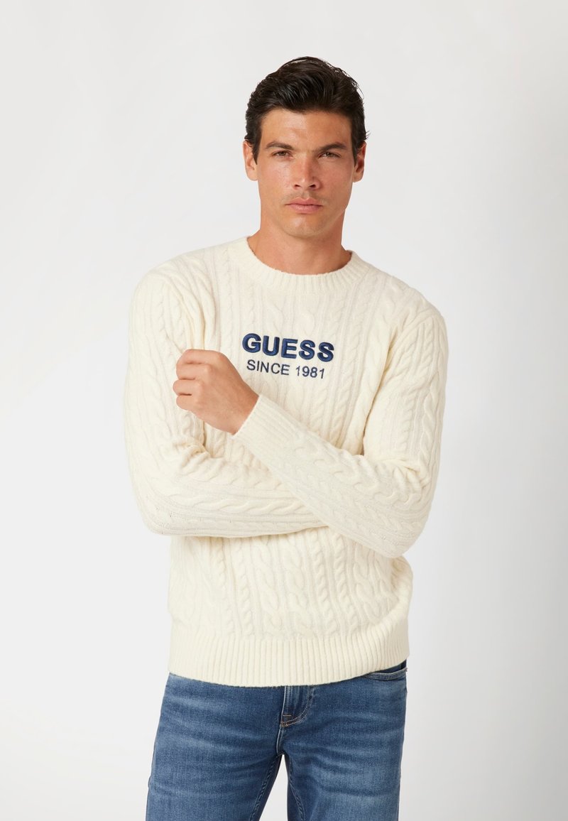 Pulover crem din tricot cu o împletitură fină, realizat dintr-un material moale. Prezintă manșete și tiv cu cusătură în relief, având un logo brodat proeminent albastru "GUESS SINCE 1981."