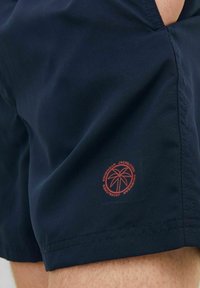 Pantaloni da bagno blu navy con design elegante, caratterizzati da un piccolo logo circolare rosso vicino all'orlo e una texture di tessuto liscia e leggera.