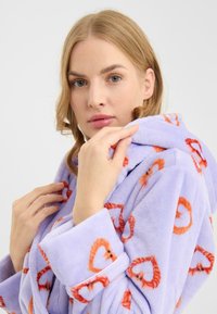 Paarse fleece badjas met een zachte textuur, voorzien van een herhalend hartpatroon in oranje. Voorzien van een capuchon en riem voor een veilige sluiting.