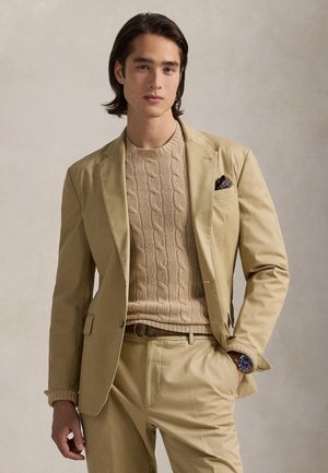 Ung mand iført beige jakkesæt og kabelstrikket sweater, med lommetørklæde og armbåndsur, stående mod en ensfarvet neutral baggrund.