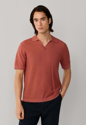 Jonge man met halflang haar, gekleed in een roodbruine korte mouwen gebreide polo en donkere broek, staand tegen een effen grijze achtergrond.