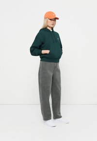 Un sweat à capuche vert avec une poche avant, associé à un jean gris ample avec un motif subtil. Le look est complété par une casquette orange vif et des baskets blanches.
