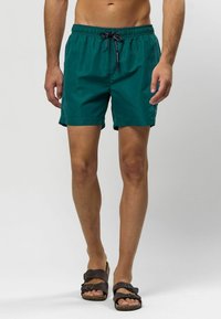 Tirkīza peldbikses ar elastīgu jostasvide un regulējamu auklu, izgatavotas no viegla auduma, kombinētas ar brūniem čībām.