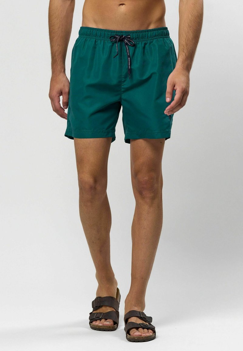 Tirkīza peldbikses ar elastīgu jostasvide un regulējamu auklu, izgatavotas no viegla auduma, kombinētas ar brūniem čībām.