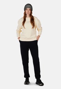 Sudadera crema con puños acanalados, combinada con pantalones de chándal negros. El modelo lleva un gorro de punto a rayas y zapatillas negras.